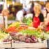 A variety of fresh vegetables displayed at a market stand with shoppers in the background. - Olive Oil Times