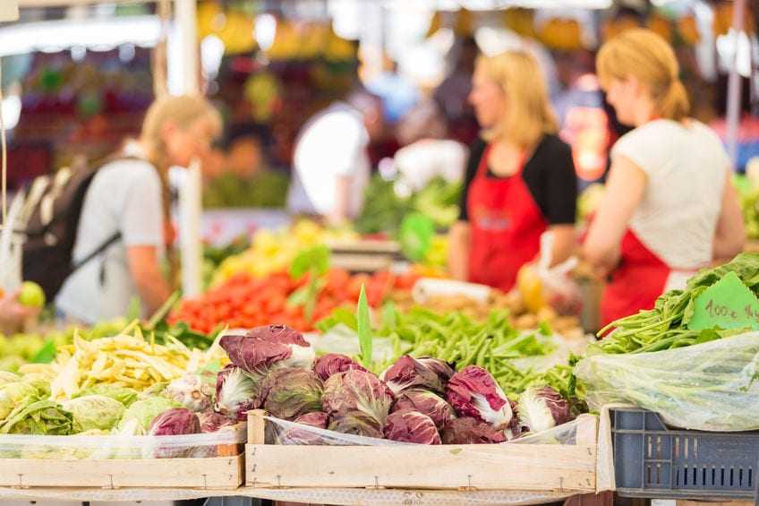 A variety of fresh vegetables displayed at a market stand with shoppers in the background. - Olive Oil Times