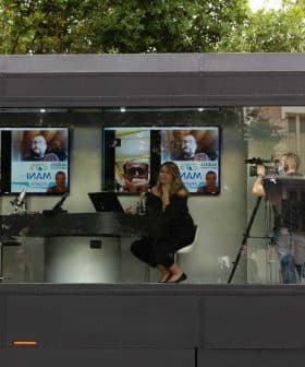 Two individuals participating in a video conference with screens displaying images in a modern setting. - Olive Oil Times