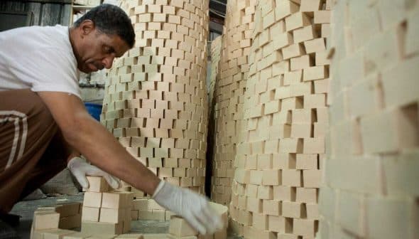 A man wearing gloves stacking rectangular soap blocks in a workshop environment. - Olive Oil Times