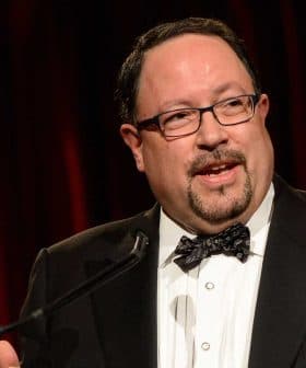 Man wearing a tuxedo and bow tie speaking at a podium during an event. - Olive Oil Times