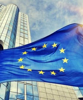 European Union flag waving in front of a modern glass building under a blue sky. - Olive Oil Times