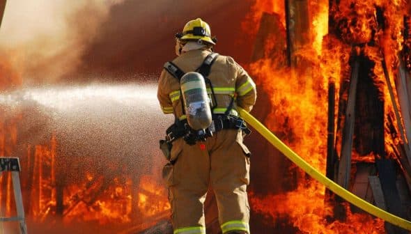 Firefighter in protective gear using a hose to extinguish flames from a burning structure. - Olive Oil Times