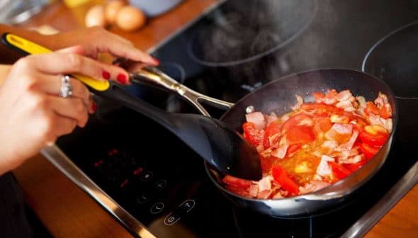 Person stirring a mixture of chopped tomatoes and bacon in a frying pan on a stovetop. - Olive Oil Times
