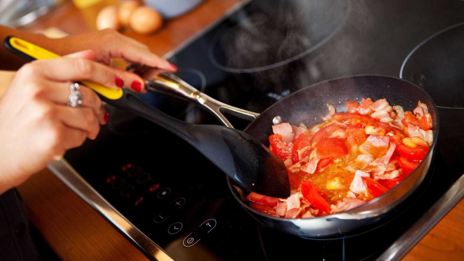 Person stirring a mixture of chopped tomatoes and bacon in a frying pan on a stovetop. - Olive Oil Times