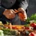 Chef peeling a carrot with various vegetables and herbs on a kitchen counter. - Olive Oil Times