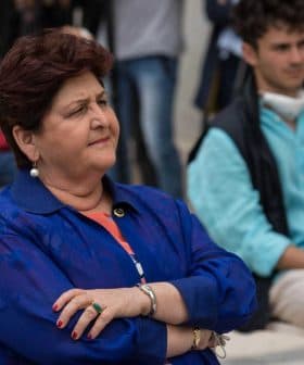 Woman with crossed arms wearing a blue jacket and earrings, seated in a group setting with other individuals in the background. - Olive Oil Times