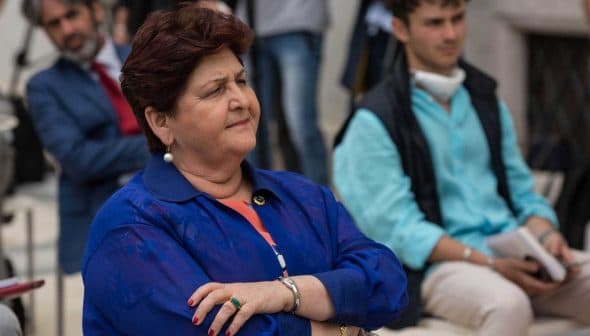 Woman with crossed arms wearing a blue jacket and earrings, seated in a group setting with other individuals in the background. - Olive Oil Times
