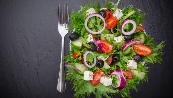 A colorful garden salad featuring mixed greens, tomatoes, cucumbers, olives, and feta cheese on a dark surface. - Olive Oil Times