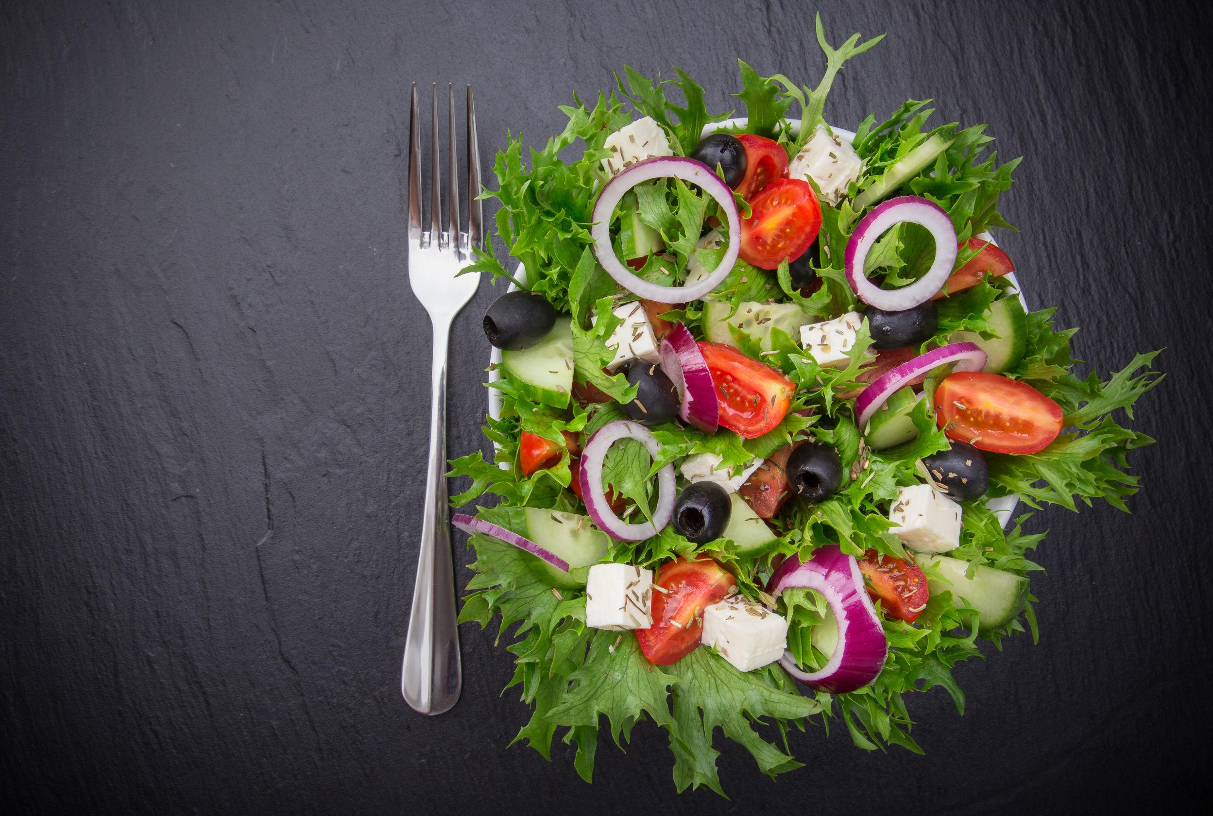 A colorful garden salad featuring mixed greens, tomatoes, cucumbers, olives, and feta cheese on a dark surface. - Olive Oil Times