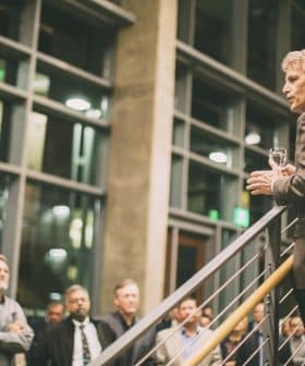 A man in a blazer speaking to an audience while holding a glass in a well-lit indoor space. - Olive Oil Times
