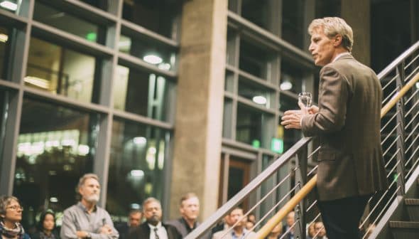 A man in a blazer speaking to an audience while holding a glass in a well-lit indoor space. - Olive Oil Times