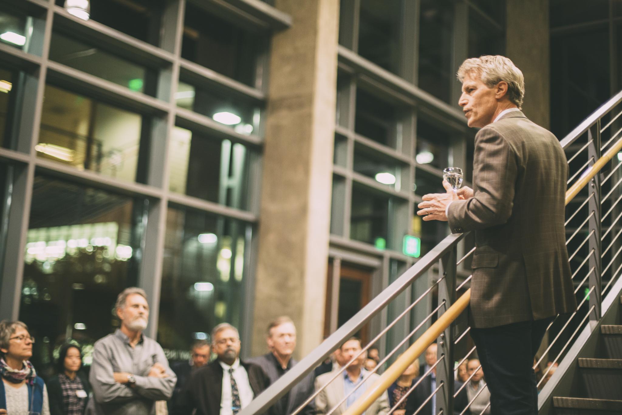 A man in a blazer speaking to an audience while holding a glass in a well-lit indoor space. - Olive Oil Times