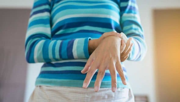 Individual wearing a striped sweater, holding their wrist with one hand while standing indoors. - Olive Oil Times