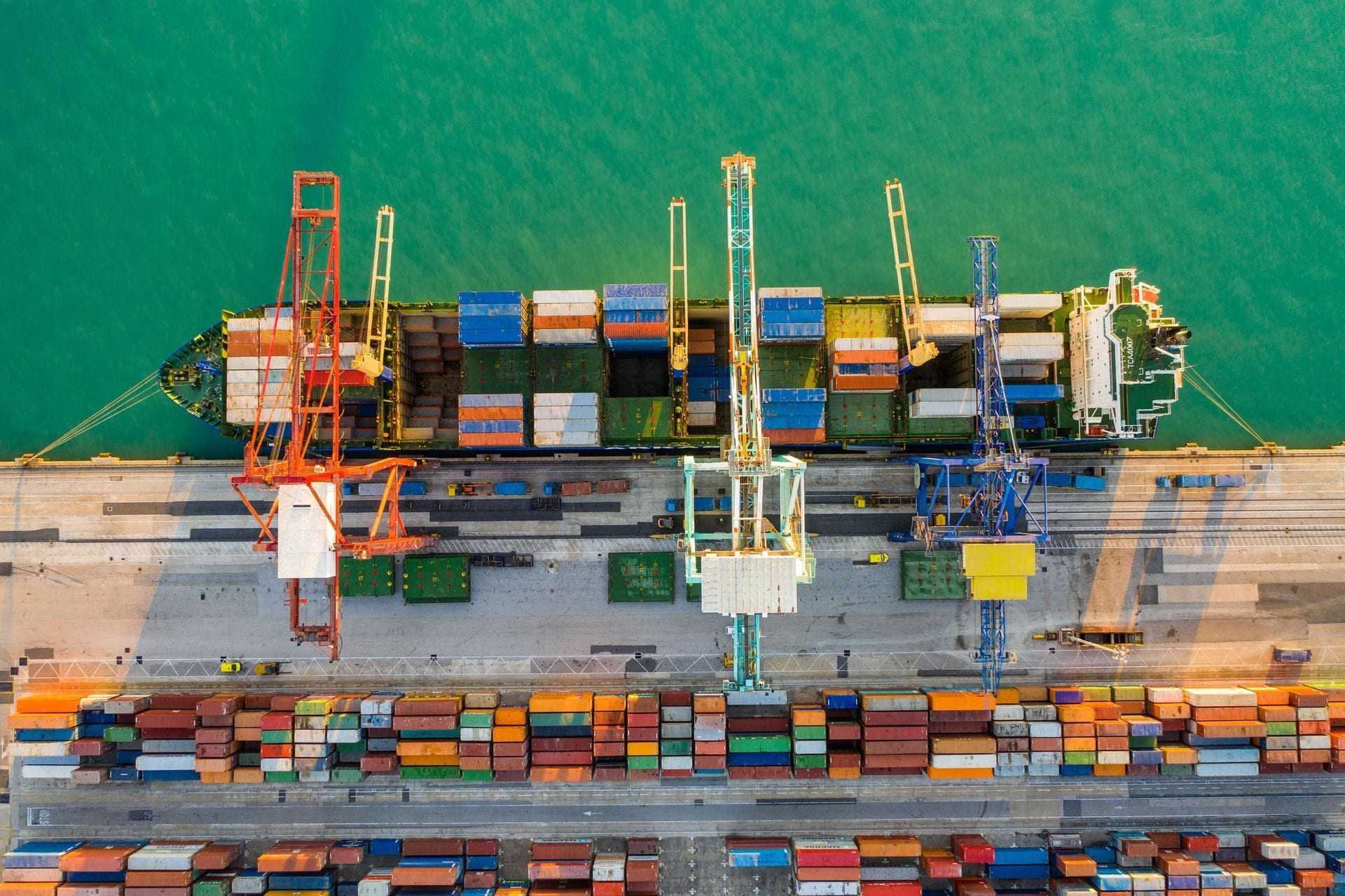 Aerial view of a cargo ship docked at a port with cranes and stacked containers. - Olive Oil Times
