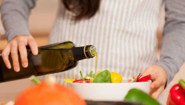 Individual pouring olive oil from a bottle into a salad bowl filled with vegetables. - Olive Oil Times