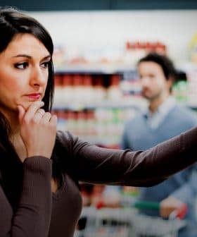 Woman with long dark hair contemplating products in a grocery store aisle while a man is in the background. - Olive Oil Times