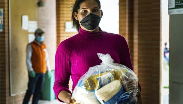 A woman wearing a pink sweater and black mask holds a bag of food items in a corridor. - Olive Oil Times