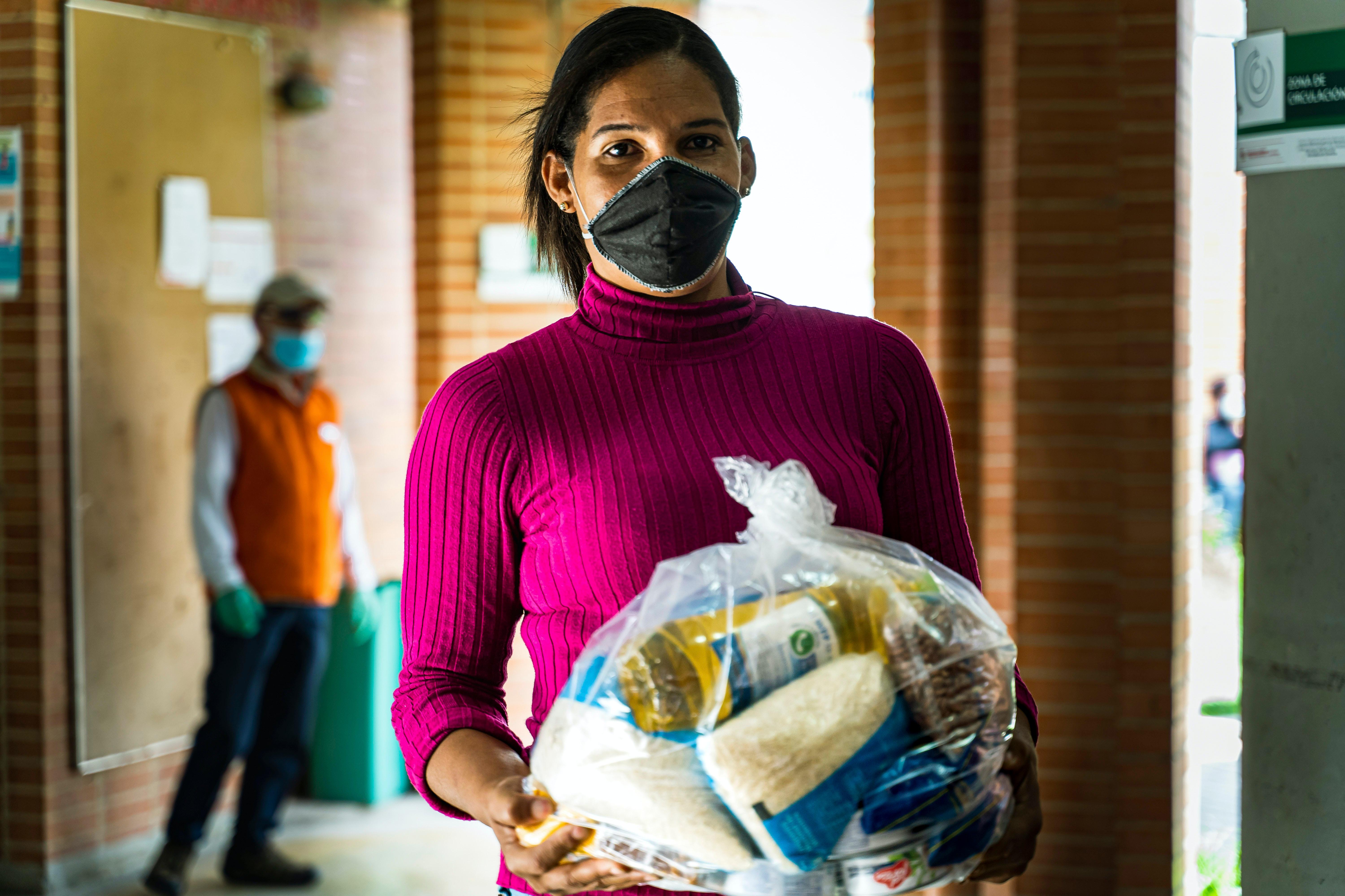 A woman wearing a pink sweater and black mask holds a bag of food items in a corridor. - Olive Oil Times
