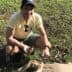 Man kneeling on grass holding olives while sitting next to a burlap sack. - Olive Oil Times