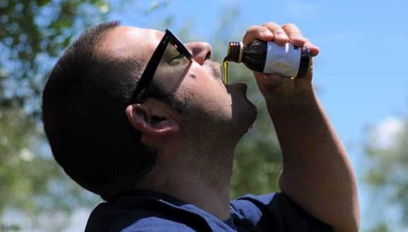 Man tilting his head back while drinking olive oil from a small bottle outdoors. - Olive Oil Times