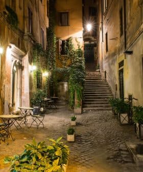 Narrow street featuring outdoor tables and potted plants, illuminated by street lamps at night. - Olive Oil Times