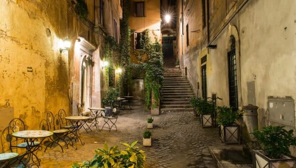 Narrow street featuring outdoor tables and potted plants, illuminated by street lamps at night. - Olive Oil Times