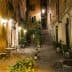Narrow street featuring outdoor tables and potted plants, illuminated by street lamps at night. - Olive Oil Times
