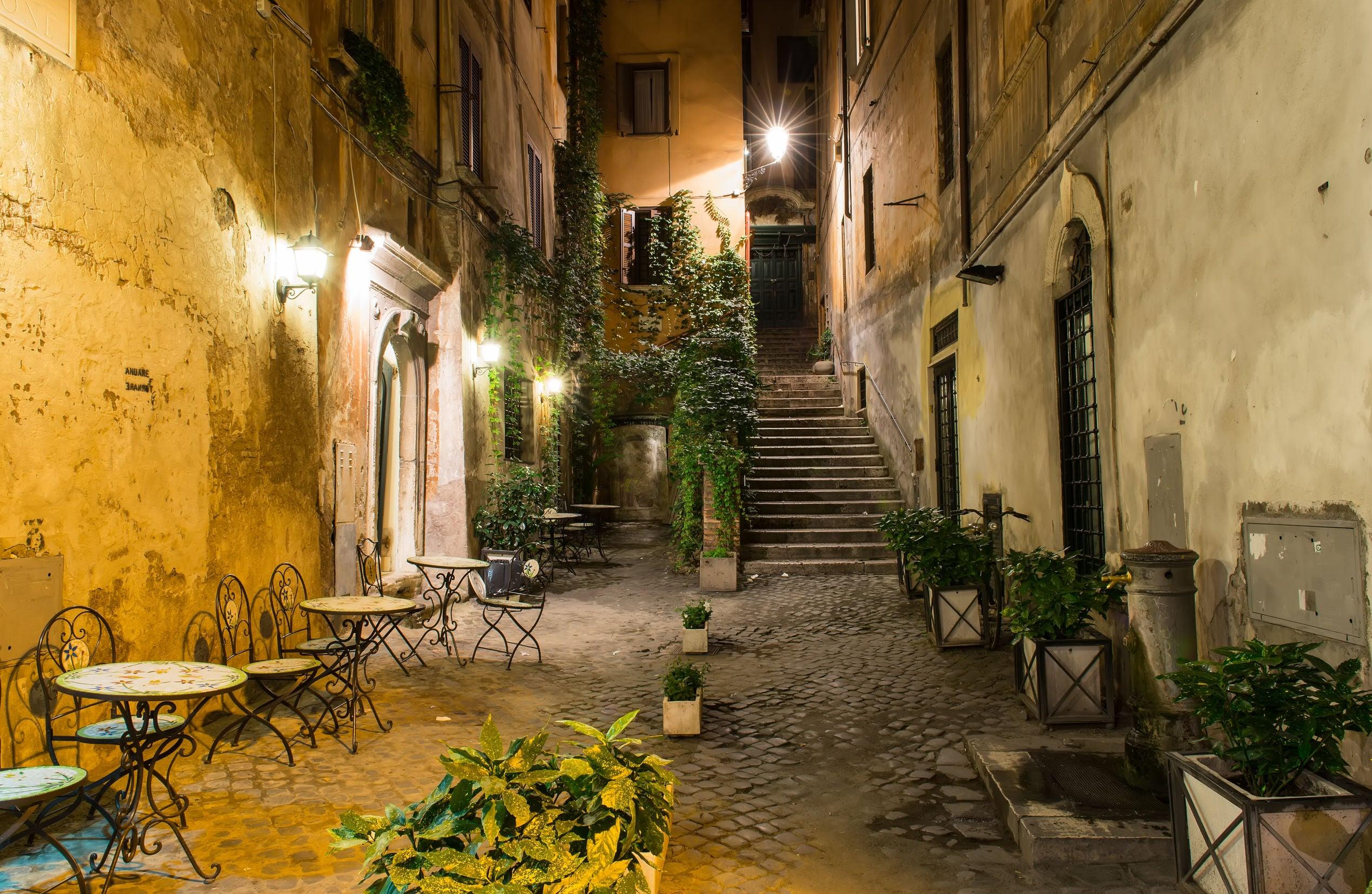Narrow street featuring outdoor tables and potted plants, illuminated by street lamps at night. - Olive Oil Times