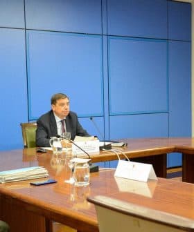Three officials seated at a conference table during a meeting, with flags in the background. - Olive Oil Times