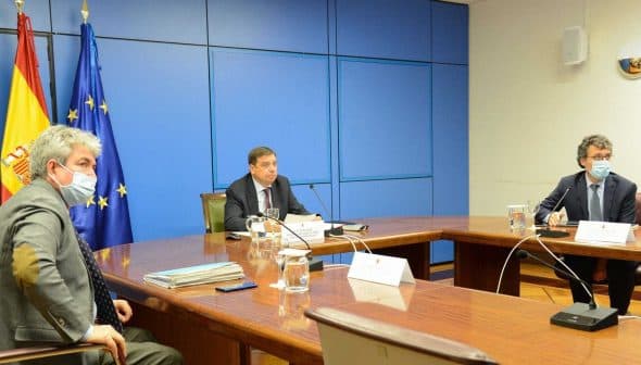 Three officials seated at a conference table during a meeting, with flags in the background. - Olive Oil Times