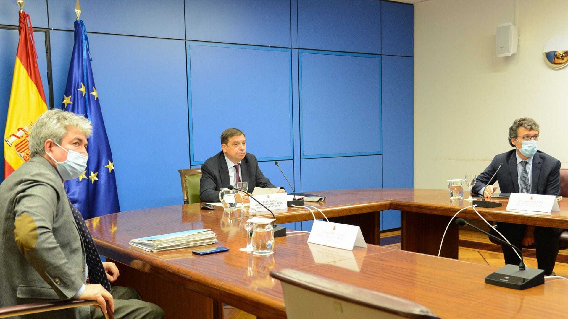 Three officials seated at a conference table during a meeting, with flags in the background. - Olive Oil Times