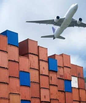 An airplane flying above a stack of colorful shipping containers at a port. - Olive Oil Times