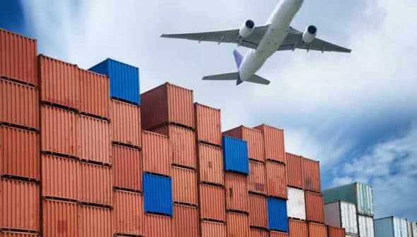 An airplane flying above a stack of colorful shipping containers at a port. - Olive Oil Times