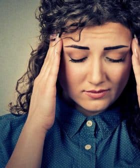 A woman with curly hair wearing a blue shirt, holding her head with both hands and closing her eyes. - Olive Oil Times