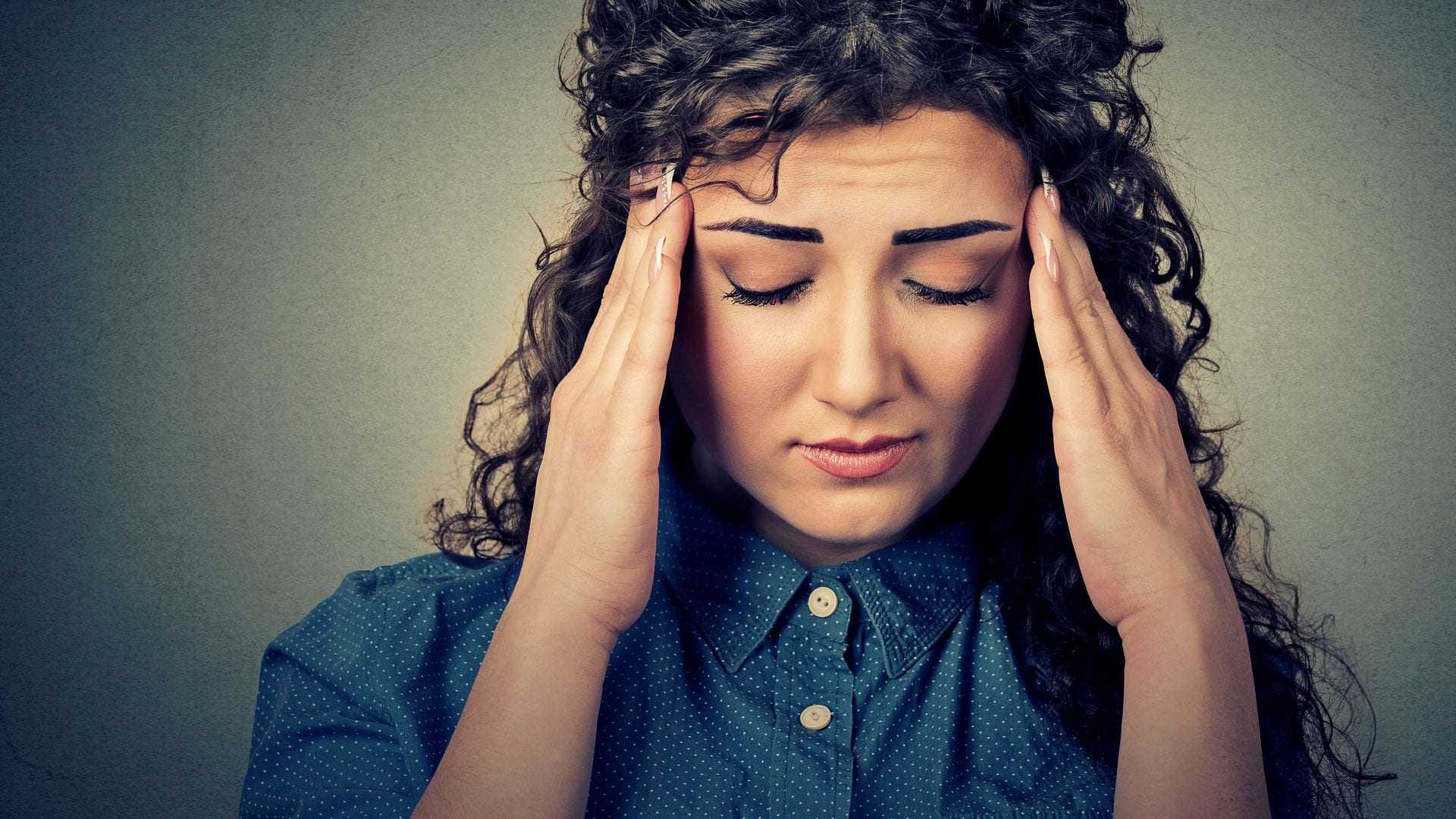 A woman with curly hair wearing a blue shirt, holding her head with both hands and closing her eyes. - Olive Oil Times