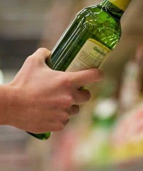 A person holding a green bottle of olive oil while reaching for another on a shelf. - Olive Oil Times
