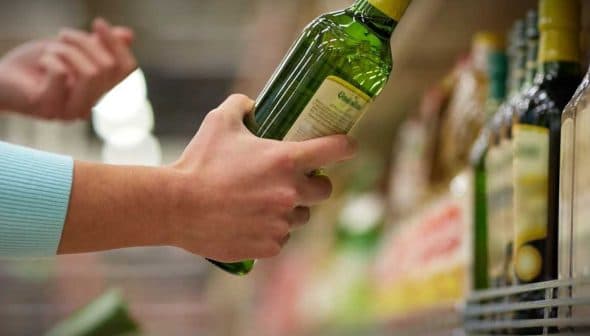 A person holding a green bottle of olive oil while reaching for another on a shelf. - Olive Oil Times