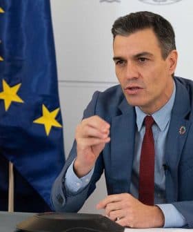 Man in a blue suit speaking during a conference with flags of Spain and the European Union in the background. - Olive Oil Times