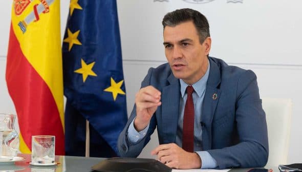 Man in a blue suit speaking during a conference with flags of Spain and the European Union in the background. - Olive Oil Times