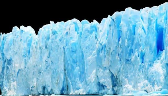 A large glacier with blue ice formations and a jagged edge against a dark background. - Olive Oil Times