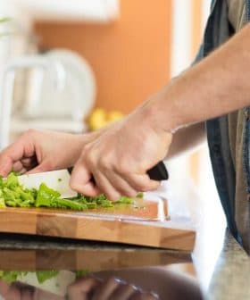A person using a knife to chop green vegetables on a wooden cutting board in a kitchen. - Olive Oil Times