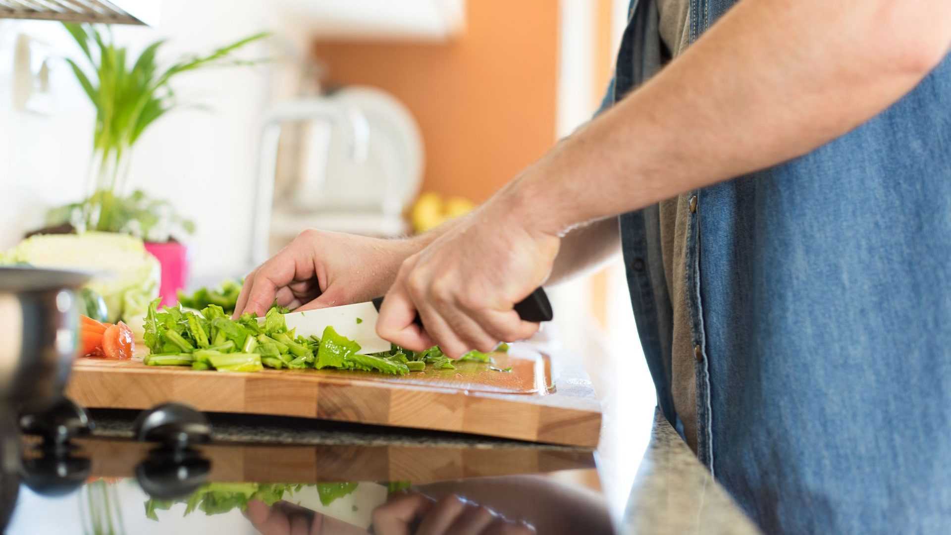 A person using a knife to chop green vegetables on a wooden cutting board in a kitchen. - Olive Oil Times