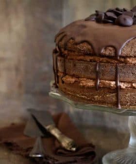 Chocolate layer cake topped with chocolate ganache and chocolate chips on a glass cake stand. - Olive Oil Times