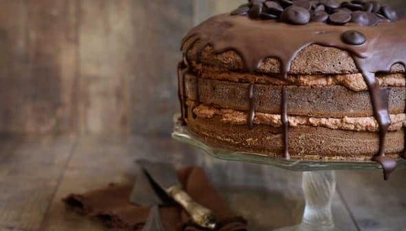 Chocolate layer cake topped with chocolate ganache and chocolate chips on a glass cake stand. - Olive Oil Times