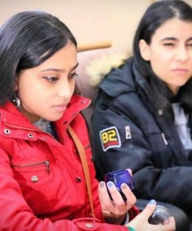 Two young women seated, one in a red jacket and the other in a black jacket, each holding a blue cup. - Olive Oil Times