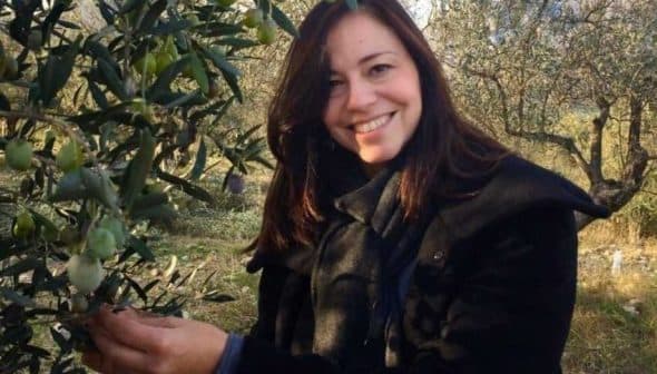 Woman smiling while picking olives from an olive tree in an outdoor setting. - Olive Oil Times