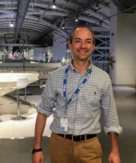 A man wearing a checked shirt and beige pants standing in a modern indoor area with a bar-like setup. - Olive Oil Times