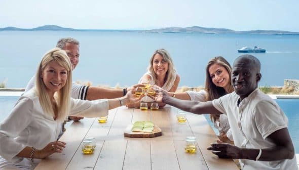 Five individuals sitting at a table, raising glasses in a toast with a scenic view of water in the background. - Olive Oil Times