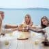 Five individuals sitting at a table, raising glasses in a toast with a scenic view of water in the background. - Olive Oil Times